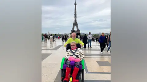 Stephan Couture Stephan in a yellow t-shirt is holding a wheelchair, with Chloe inside it. She is wearing a black and white striped top, blue legging and bright red boots. They are both looking towards the camera with the Eiffel Tower in the background. A number of people are also walking around an ornate cream, black and white tiled plaza behind Stephan and Chloe