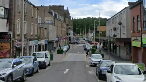 Google A high street with shops on each side. Cars are parked on either side of the road.