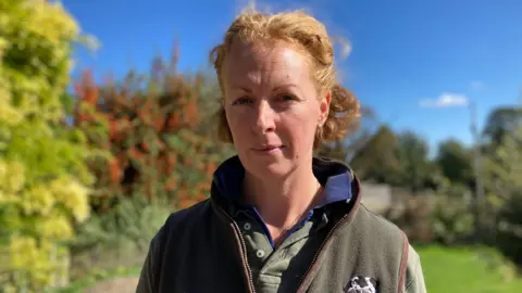 Farmer with red hair wearing green top and stood outside near trees. There is a blue sky behind her.