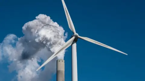 Getty Images Wind turbine in front of smoking chimney