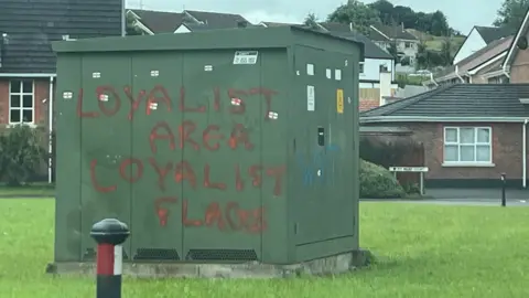 Shows graffiti on an electricity facility saying "LOYALIST AREA LOYALIST FLAGS"