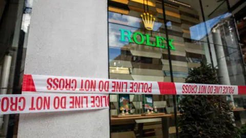 A red and white police tape outside the Rolex store on Knightsbridge which is shown with green writing and golden crown behind glass