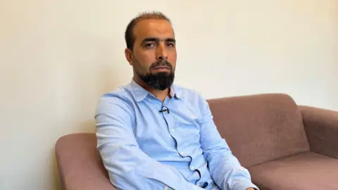 Man of Afghan origin in blue shirt short black hair sat on brown sofa