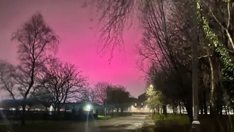 Dave T Callacher A bright pink glow lights up the whole night sky in shades of pink and mauve on a residential street-lit road.