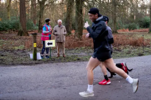 Aaron Chown/PA Two runners and a dog on a metalled road at Sandringham Parkrun. King Charles and a race volunteer are standing at the edge of the road, with woodland behind them.