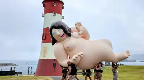 A naked inflatable character being carried by five strong people on the Hoe in Plymouth. Smeaton's Tower, a red and white striped lighthouse, is in the background. The sea can be seen in the left of the image in the background.