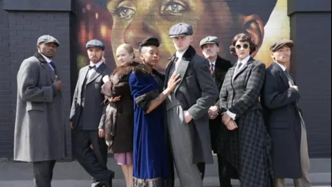 The cast of the Peaky Blinders stage show, lined up in front of a mural of the actor Cillian Murphy as Thomas Shelby.