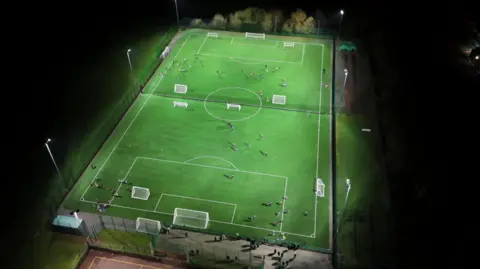 A drone shot of an artificial all weather surface football pitch which is lit by flood lights. Players are training on the pitch. 