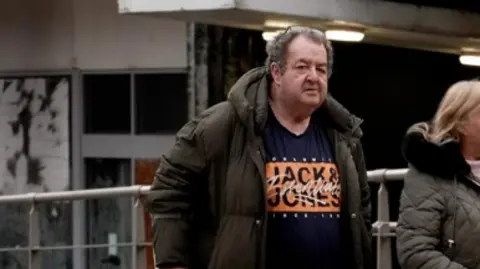 Gerard Woodhouse, wearing a raincoat and a Jack Jones t-shirt and using a walking stick, walks into Liverpool Magistrates Court. 