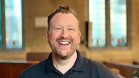 A man with brown hair and a beard smiles with his teeth at the camera. He wears a navy blue polo shirt and stands inside a church.