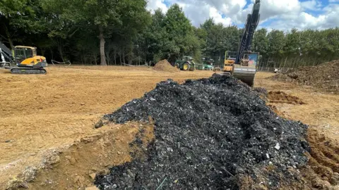 Environment Agency A large mound of dark waste is in a pile in front of a digger.