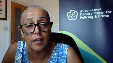 A woman with short hair and wearing glasses sits in front of a blue and green banner saying "Alison Lowe Deputy Mayor for Policing and Crime"