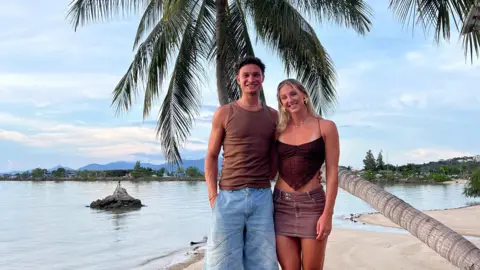 Joel Clarke-Khan The couple stand in front of a palm tree on a beech next to the sea. Joel has short dark hair, wears a sleeveless brown top and light brown denim shorts. Molly has a dark red top and a faded red skirt. They have arms around each other.
