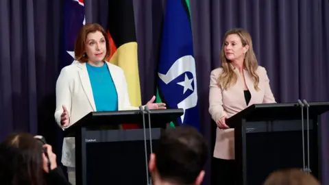 Getty Images Anika Wells, wearing a blue top and white jacket, speaks at a lecturn while a blond woman in a pink jacket watches on