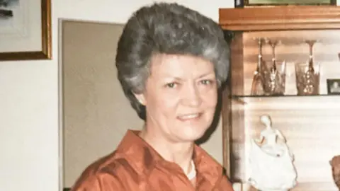 Margaret Joy Todd with short grey hair, wearing a burnt orange coloured shirt, smiling and standing in front of a a wooden cabinet displaying a porcelain figurine and crystal glasses