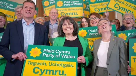 PA Media A group of Plaid Cymru members and leaders holding yellow and green banners which say Plaid Cymru, Party of Wales, and "Tegwch, Uchelgais. Cymru", smiling to the camera.