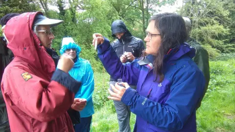 River Foss Society A small group of people in waterproof jackets stand in a wooded area during wet weather. One person holds a small container and tests water.