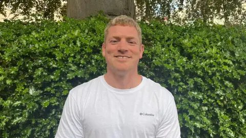 Jason standing in front of a tree and bush wearing a white T-shirt and smiling at the camera