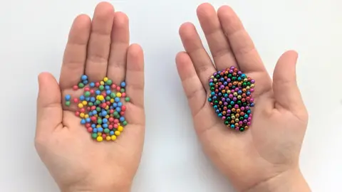 two hands holding tiny multi-coloured ball bearings on the left is some which are sweets and on the right is some which are a toy
