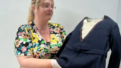 Leeds City Council A woman wearing glasses and a floral dress holds up a children's navy blue sailor suit top.