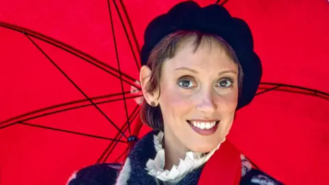 Shelley Duvall smiling in front of a bright red umbrella