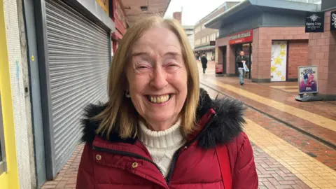 Photograph of 75-year-old Wyn Bailey from Wythenshawe. She is pictured at the Civic Centre in the town. Smiling for the camera and wearing a red coat over a cream jumper, Wyn has blonde, collar-length hair.
