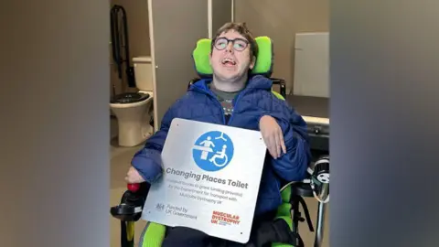 Zack Kerr with brown hair and glasses wearing a blue coat and grey patterned jumper smiling holding a Changing Places toilets plaque 