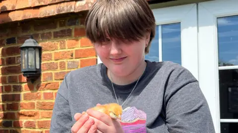 A young woman carefully holds a week-old yellow chick in both her hands. She is smiling at the camera and has short dark hair and a black T-shirt.