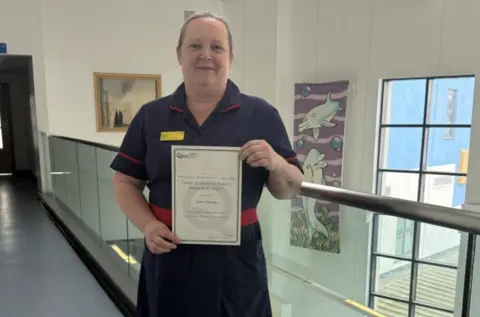 The Royal Wolverhampton NHS Trust A woman in uniform is holding a piece of paper including the words Dame Elizabeth Fradd Memorial Prize. She is standing in a glass-walled corridor and a window is to her right.