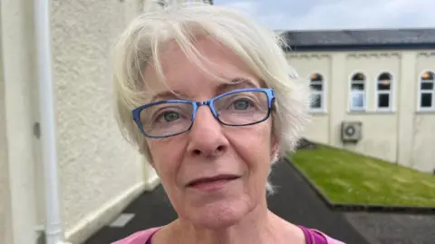 A white haired woman has blue glasses on. She wears pink top. Behind her is a cream coloured building.