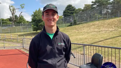 Will Alonso wearing a dark grey cap and a green T-shirt underneath a black zip up sweatshirt. He is smiling at the camera, standing on the orange-red track at the stadium. Beside him are some of the adaptable bikes. 