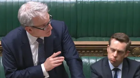 Damien Hinds with short white hair and glasses wearing a blue shirt and standing up in the House of Commons