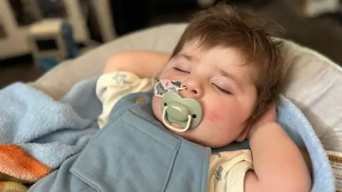 Jack is asleep with his hands behind his head. He has brown hair, is in a blue baby grow and has a dummy in is mouth and a feeding tube going up his nose.