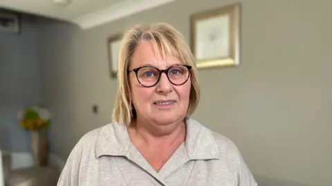 Kate Bradbrook/BBC A woman with shoulder length blonde hair smiles at the camera. She is standing in a living room and is wearing black glasses with a grey top on.