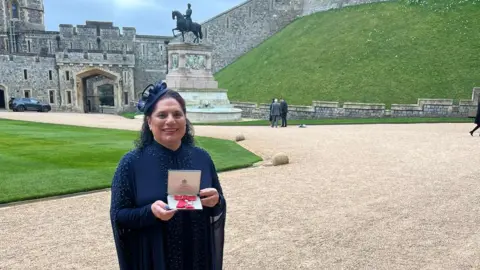 Razia Hadait A woman in blue is holding her MBE, which is facing the camera. She is smiling and on the left side of the photo, with a large walkway and grass behind her.