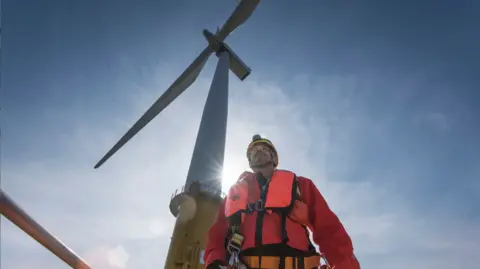 Male worker in florescent orange workwear and yellow helmet stands in front of an offshore wind turbine