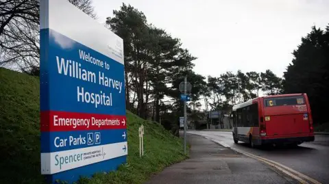 A sign outside the William Harvey Hospital points the way to the emergency departments, car parks and other sites. There is a bus on the road which has double yellow lines. The sign stands on a grass slope.
