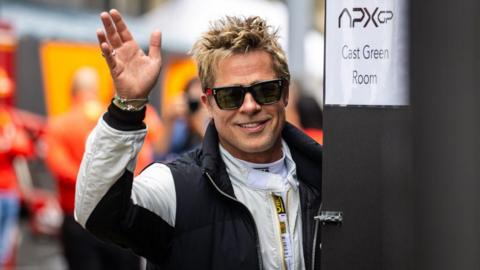 Brad Pitt, wearing dark glasses and a racing suit, waving at fans at Silverstone