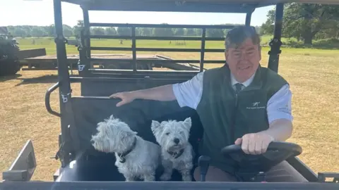 Colin Rayner is driving a quad bike with his two West Highland terrier dogs sitting to his right hand side as he holds the wheel. Grass fields can be seen behind them.
