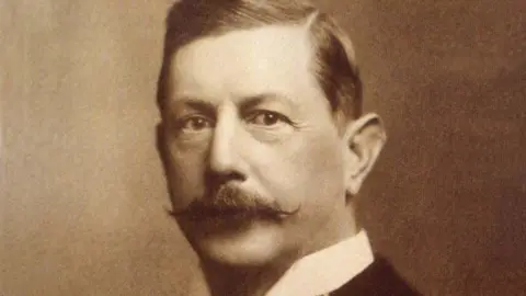 A sepia head shot of Edward Stocks Massey. He is wearing a smart black coat with a white stand-up collar and his hair is neat and slicked back with a side parting and a moustache that is neatly turned up at the ends