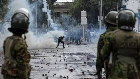 Security officers in uniform (foreground) facing a protester in the background who ducks a teargas canister on a street littered with debris and smoke
