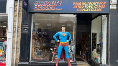 BBC A full-sized, roughly 7-foot-tall statue of Superman stands outside on the high-street of a storefront of a retro toy shop named "Curiosity Classics”. The statue stands in a heroic, authoritative stance with hands planted firmly on his hips. He wears the classic suit with a cape. The statue stands on a grey, stone-textured rectangular block featuring the raised inscription "MAN OF STEEL."