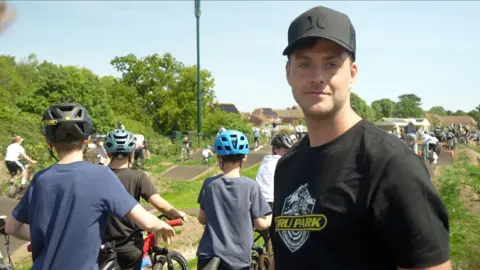 Connor Bennett/BBC Jono Jones is looking at the camera wearing a black T-shirt and baseball cap. Children with bicycles can be seen behind him.