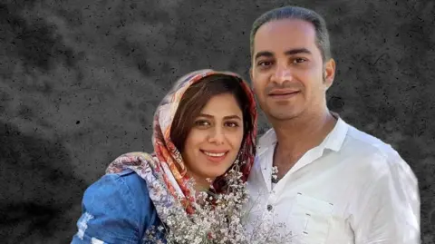 Mansoureh Heydari and her husband, Behrouz Mansouri, smiling and embracing as they look at the camera. Mansoureh wears a headscarf and holds a bouquet of flowers, Behrouz wears a white shirt.