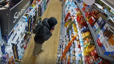Cambridgeshire Police Man with a dark puffer jacket and hoodie puts chocolate into his jacket in a convenience store aisle.