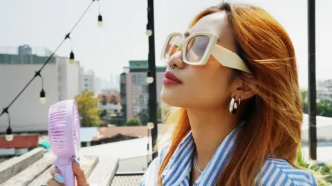 Getty Images An asian woman with red dyed hair wearing sunglasses holds a pink mini-fan