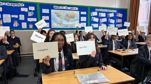 Secondary school children sitting in a Geography classroom. There are two pupils per desk and each pupil is holding up a white board with the word 'metaphor' on it. Behind the children there is a map of the world on a blue background. 