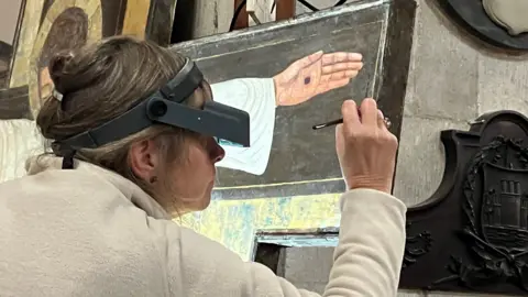 Woman with her hair tied up and wearing a magnifier on her head in order to see a close up of the painting she is restoring. She has a paintbrush in her hands and is working on the side of the painting. You can see part of the painting which depicts Jesus Christ's hand and a black dot which represents the crucifixion