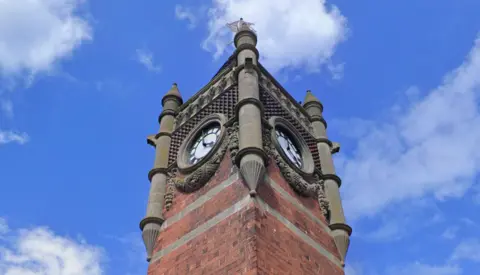 A picture angled up at the King Edward VII Memorial Clock Tower focused on the top part which has two clockfaces visible with ornate stone carvings surrounding them.