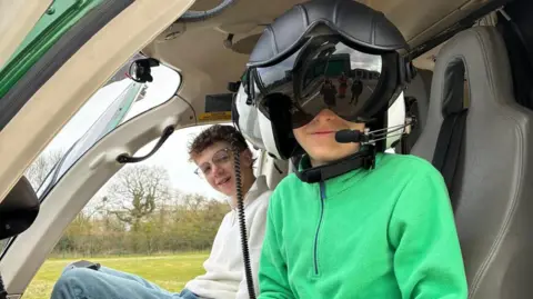 Family photo Two teenage boys smile from the cockpit of a helicopter on the ground. The one in the foreground wears a large pilots helmet with the visor down.